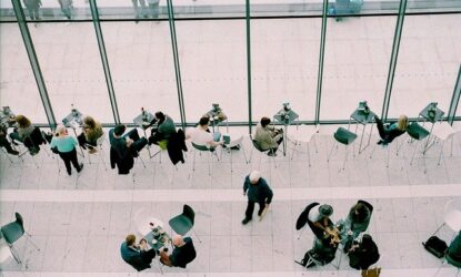 Zdjęcie przedstawia grupę osób skupionych wokół stolików, stojących, siedzących. Pomieszczenie w jakim się znajdują to prawdopodobnie sala konferencyjna. Zdjęcie wykonano z góry, z tzw. lotu ptaka.