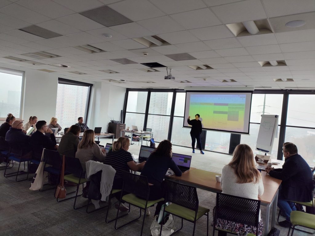 Zdjęcie przedstawia grupę osób podczas szkolenia. Uczestnicy siedzą przy stole, skierowani twarzami do prowadzącej szkolenie stojącej przy tablicy. 