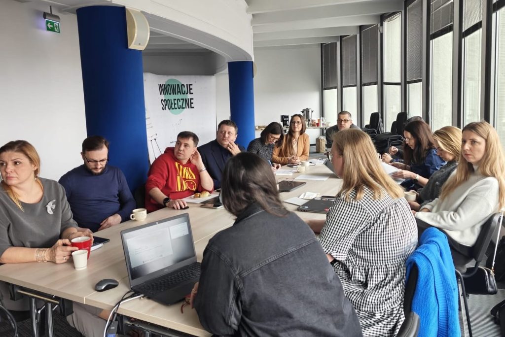 Zdjęcie ze spotkania grupy osób. Wszyscy siedzą przy stole. Wzrok większości osób jest skierowana w jedną stronę. Osoby te obserwują prezentację.