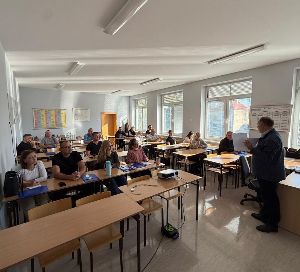 Szkolenie - kilkanaście osób siedzi przy biurkach, w ławkach. Zwrócona do nich przodem osoba - mężczyzna prowadzi szkolenie, przemawia do zebranych uczestników.